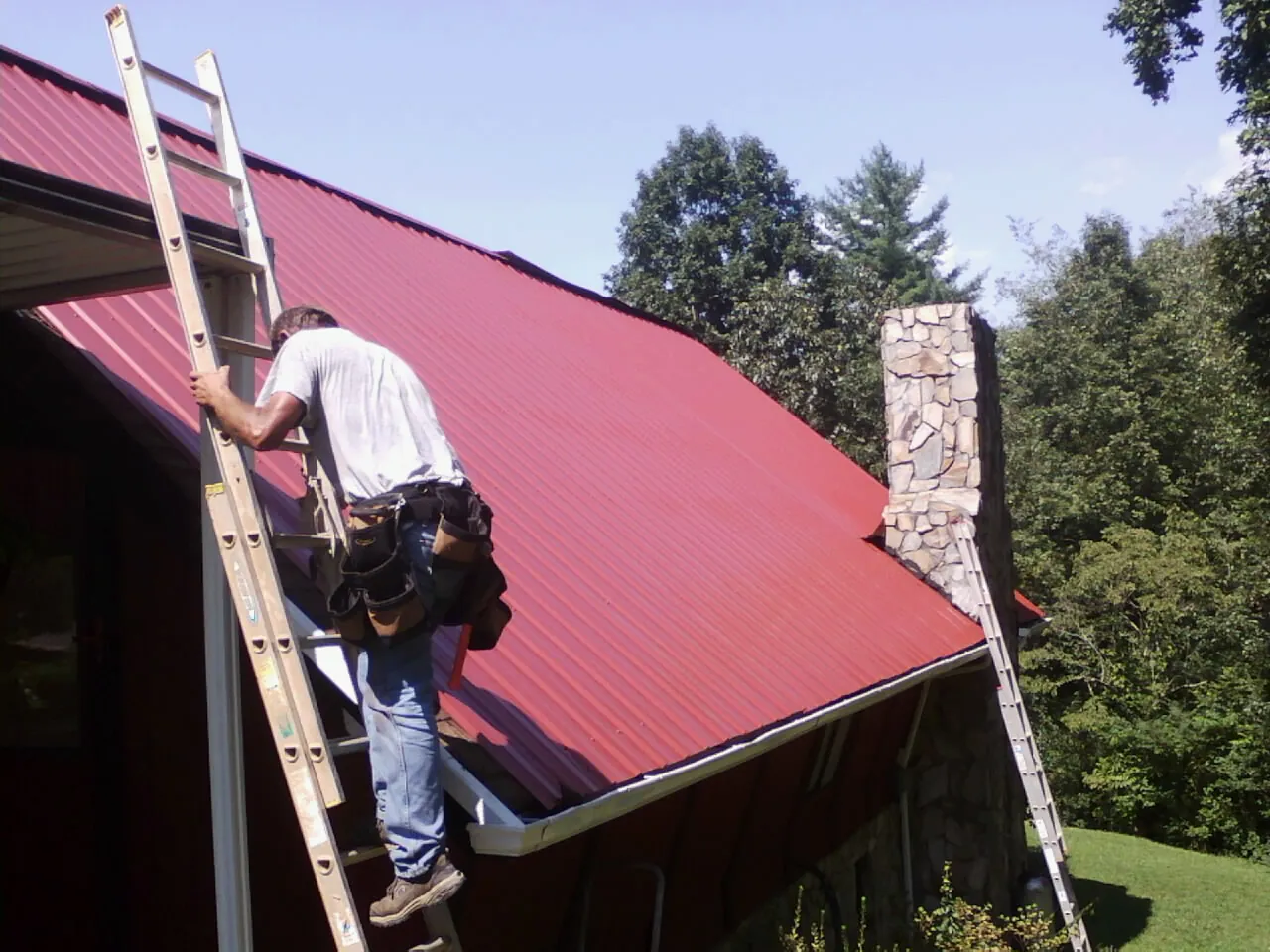 Roofer on a ladder beside a red metal roof and stone chimney — Soffit & Fascia Repair by Roofing Brothers in Leetsdale
